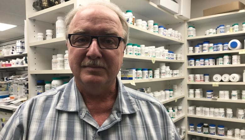 A man with a mustache and glasses stands in front of the shelves of a pharmacy.